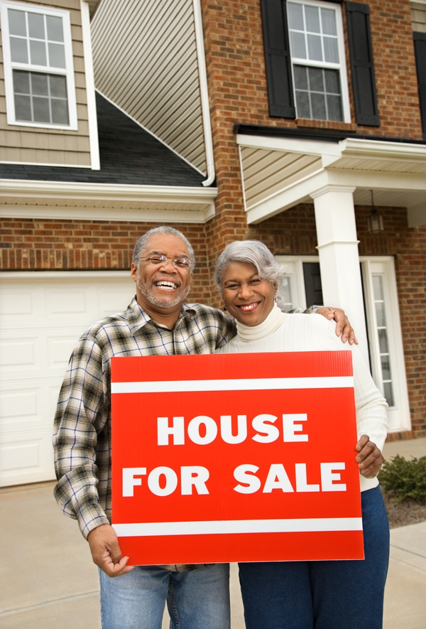 Two seniors standing behind a House for Sale sign.